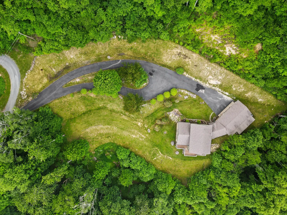 A serene exterior perspective featuring the home, a winding driveway, and lush greenery. The porch is visible, inviting thoughts of relaxed mornings soaking in natural surroundings. The gentle curve of the drive and layers of trees suggest privacy within a gated community.