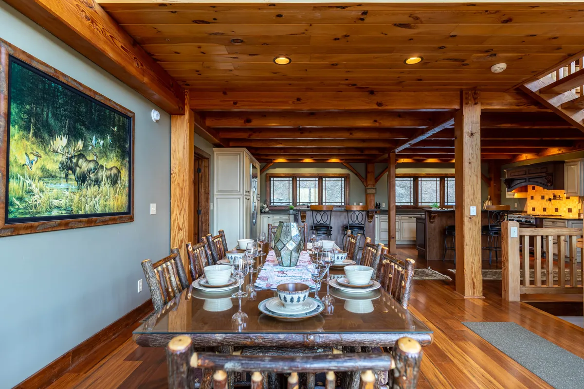 A warm, communal interior showcasing a large wooden dining table beneath rustic beams, with a painting of a moose on the wall and a view into the adjoining kitchen. The space evokes the sense of togetherness, perfect for family meals, celebrations, and group gatherings.