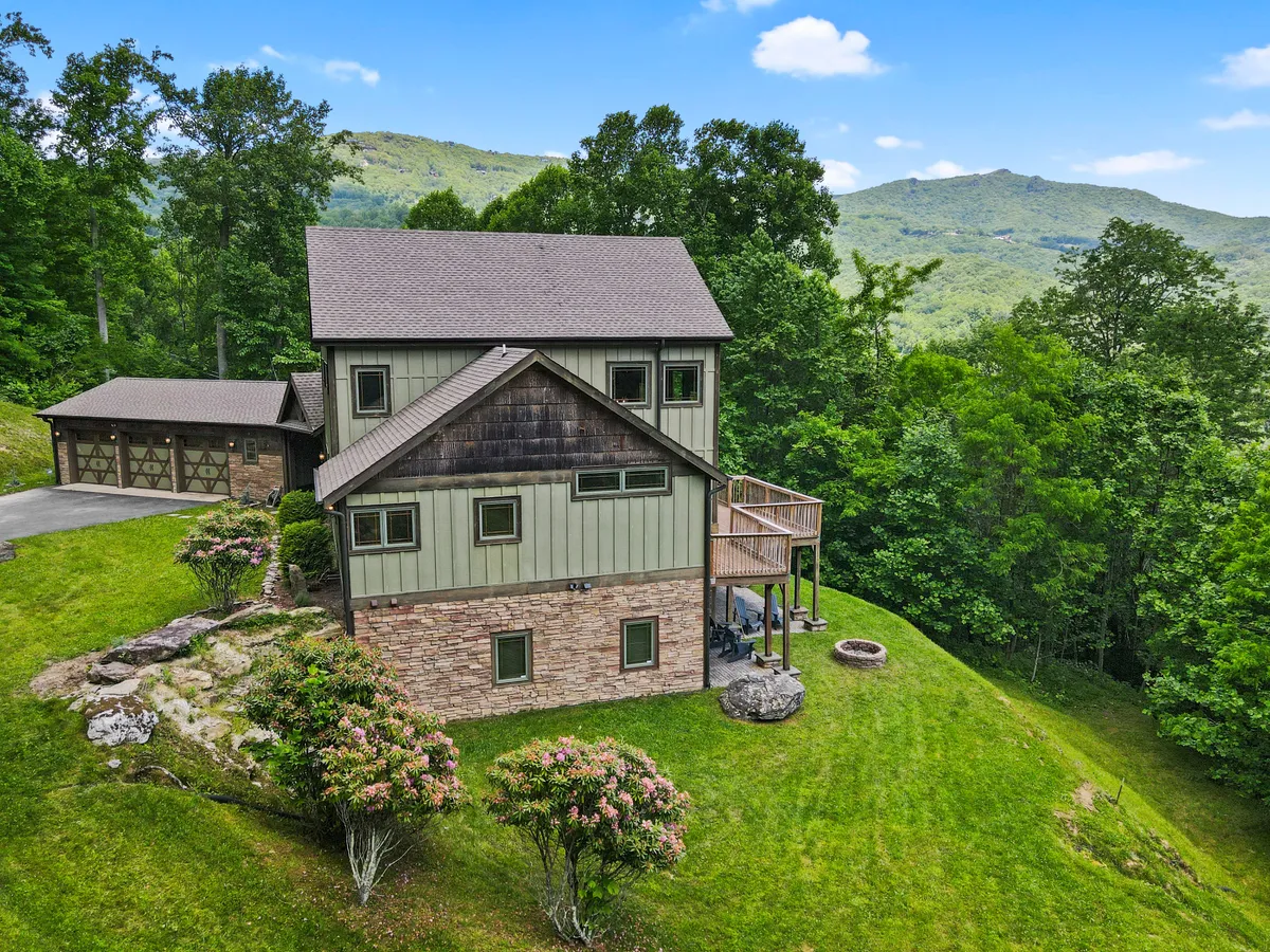 A broad exterior view of the modern house that showcases a stone fire pit, the inviting deck, and the natural landscape of surrounding trees. The scene suggests spaces meant for both gathering and relaxation, especially in the evenings.