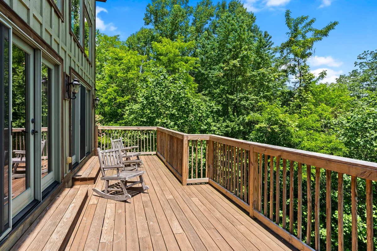A wide wooden deck surrounded by lush greenery features classic rocking chairs, creating a cozy spot to relax and enjoy the view. The natural backdrop and inviting seating encourage unwinding outdoors while keeping an eye on children playing in the yard.