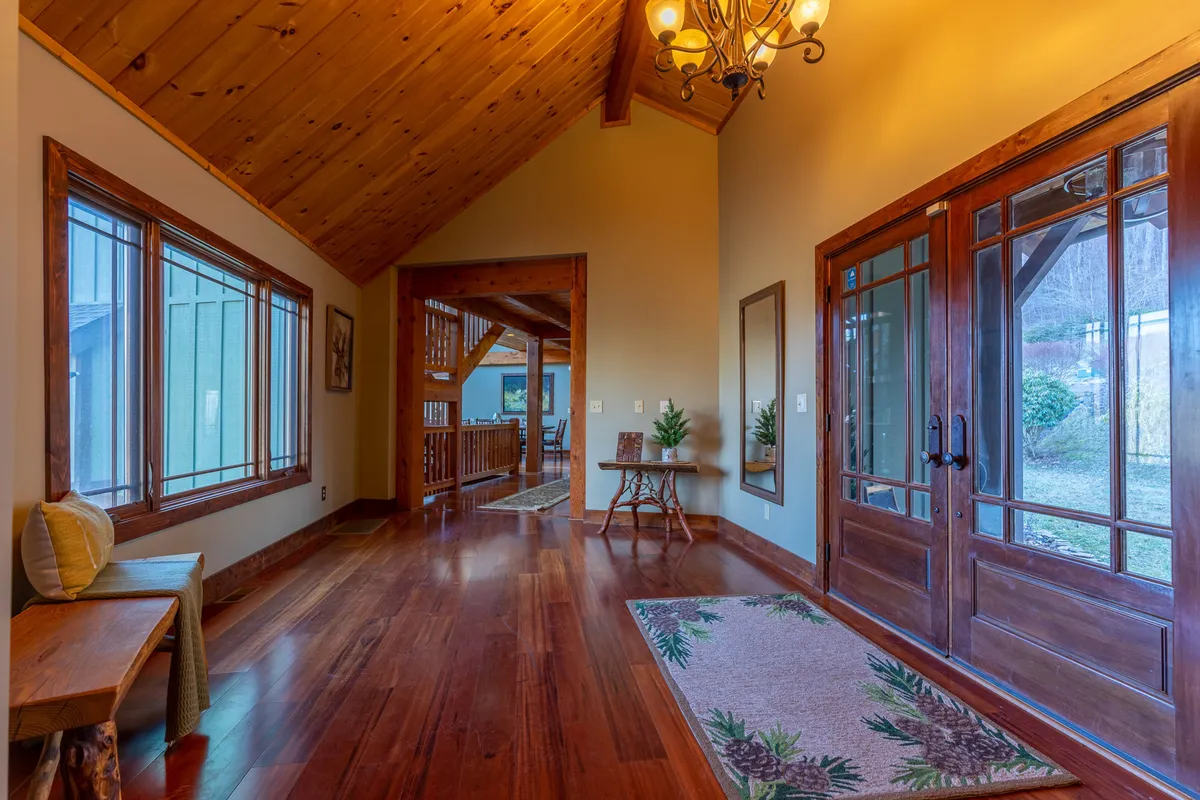 A welcoming entryway with a sturdy wooden front door, a warm chandelier overhead, and sunlight pouring in through large windows. The inviting space features a decorative rug and wood flooring, hinting at the warmth and hospitality within. This area feels like the natural threshold where friends arrive before their mountain adventure begins.