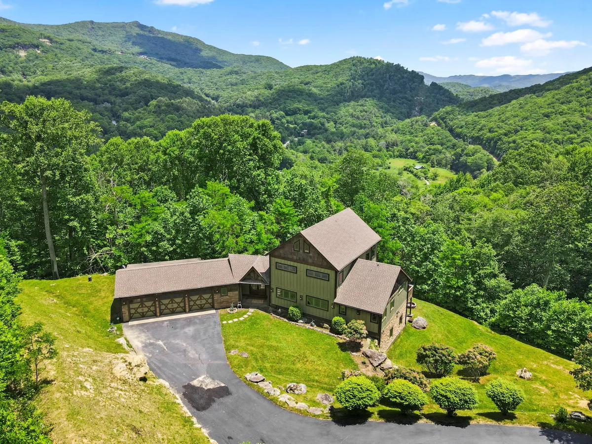 The hero image features the striking exterior of a luxury mountain home set against a breathtaking backdrop of rolling peaks and lush greenery. The house stands proudly on beautifully landscaped grounds, surrounded by mature trees and vibrant lawns, with the majestic Blue Ridge Mountains stretching into the distance. The setting is serene and private, perfect for outdoor gatherings or simply soaking in the natural beauty.