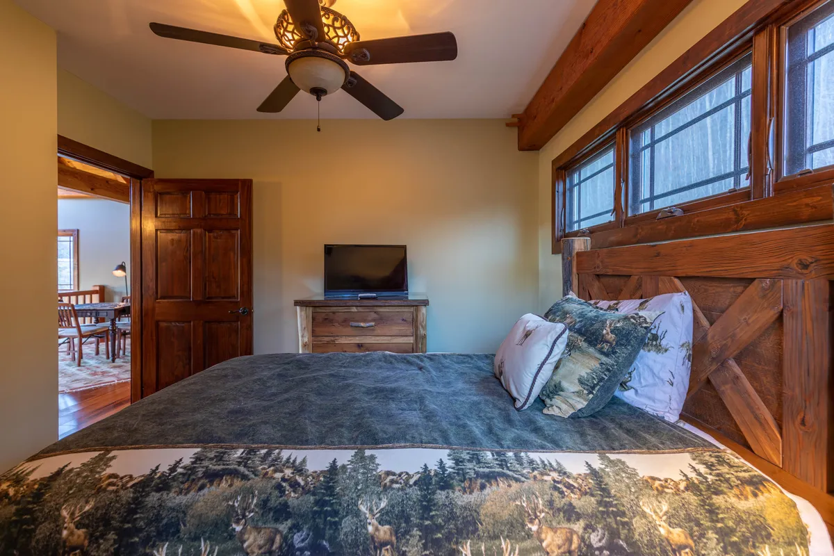A tranquil bedroom featuring a bed with wildlife-themed bedding, a sturdy wooden headboard, a TV on a dresser, and a large window bringing in natural light. The room blends rustic charm with modern comfort, providing a cozy retreat for relaxation and entertainment.