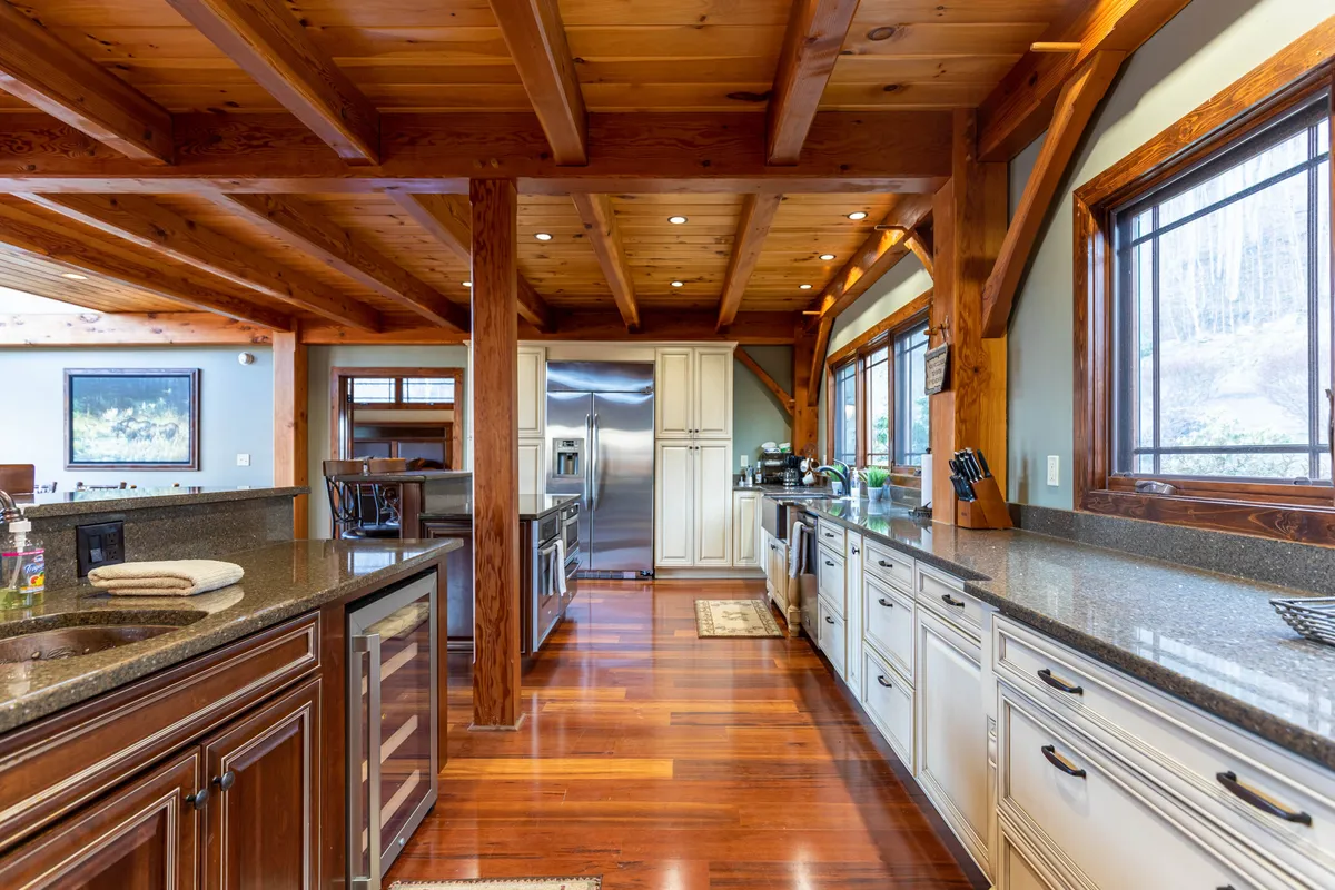 A beautifully appointed kitchen with granite countertops, exposed wooden beams, stainless steel appliances, and broad windows letting in natural light. The space is designed for group cooking and entertaining, with enough room for several people to participate.