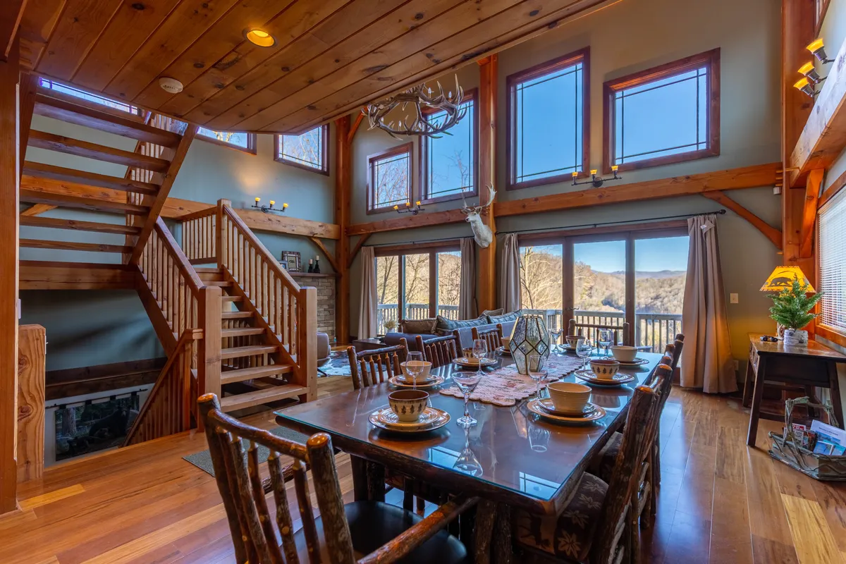 An expansive, open interior with a large dining table perfect for group meals or game nights. Expansive windows reveal natural light and mountain views, while the adjacent cozy seating area and decorative antlers evoke a rustic, social atmosphere. The staircase in the background connects this main space to other areas, suggesting movement and communal activity.